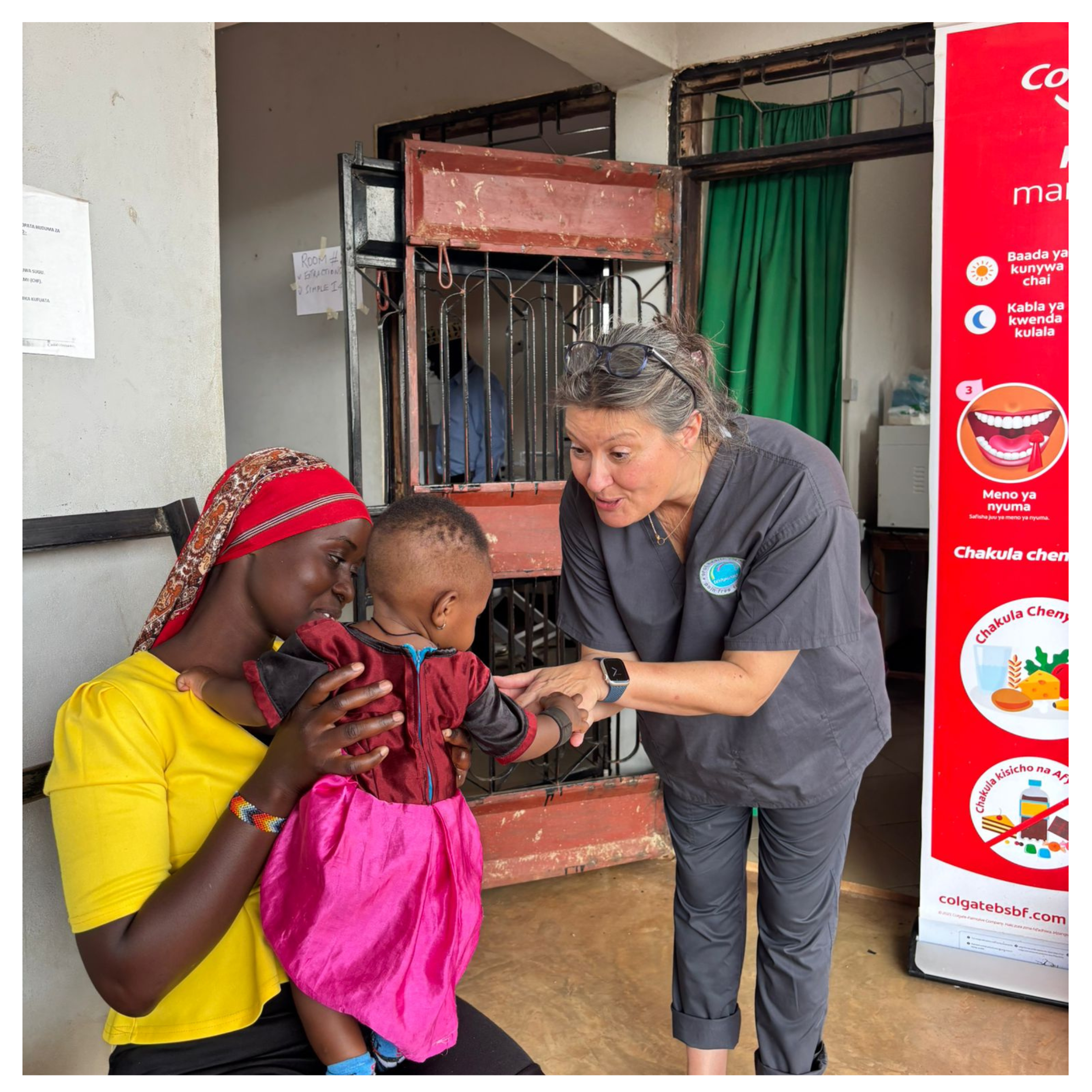 A healthcare volunteer in grey scrubs smiles and gently interacts with a baby held by a mother in a bright yellow top and red headscarf inside a clinic setting in Tanzania.