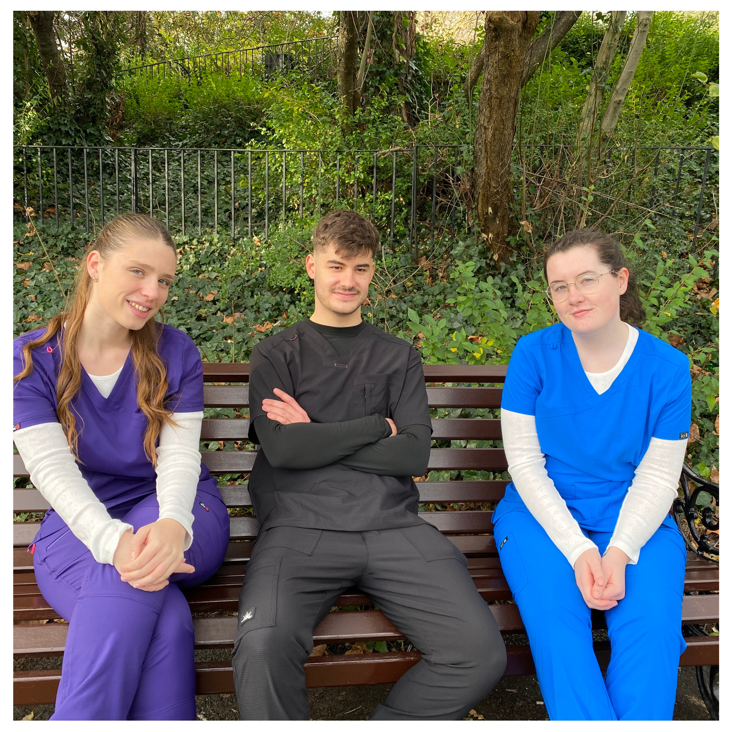 Three physios wearing physio uniforms in purple, black and blue sit on a wooden bench outdoors, surrounded by greenery, creating a relaxed and professional team atmosphere.